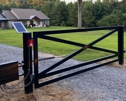 Black metal gate with solar panel and house in the background. Gravel driveway, green grass.