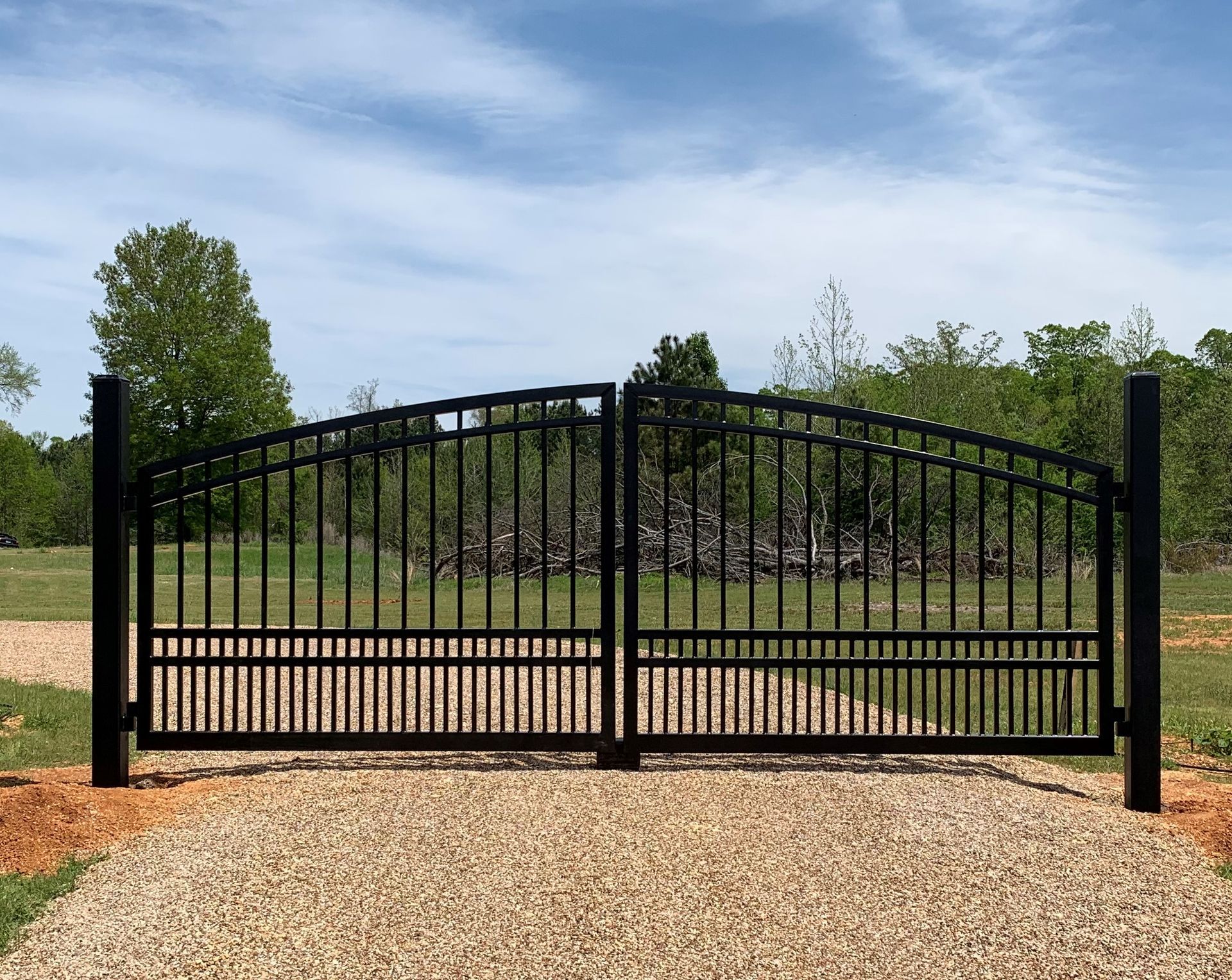 Black metal driveway gate on gravel path, set against a field and sky with clouds.