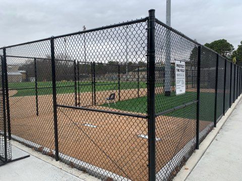 Black chain-link fence surrounds a small baseball field with tan infield and green turf.