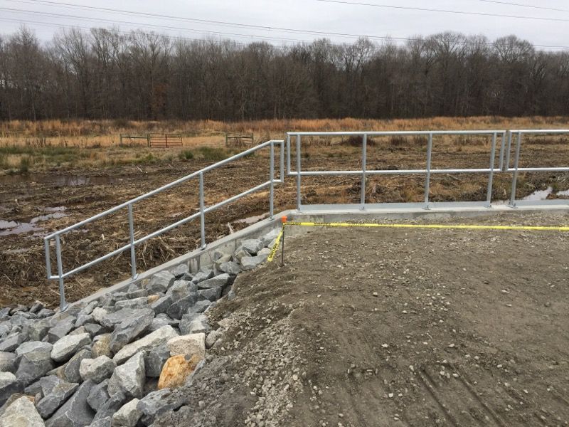 Metal railing on a concrete barrier and rocky embankment, leading to a dirt and gravel path with a field and trees in the background.