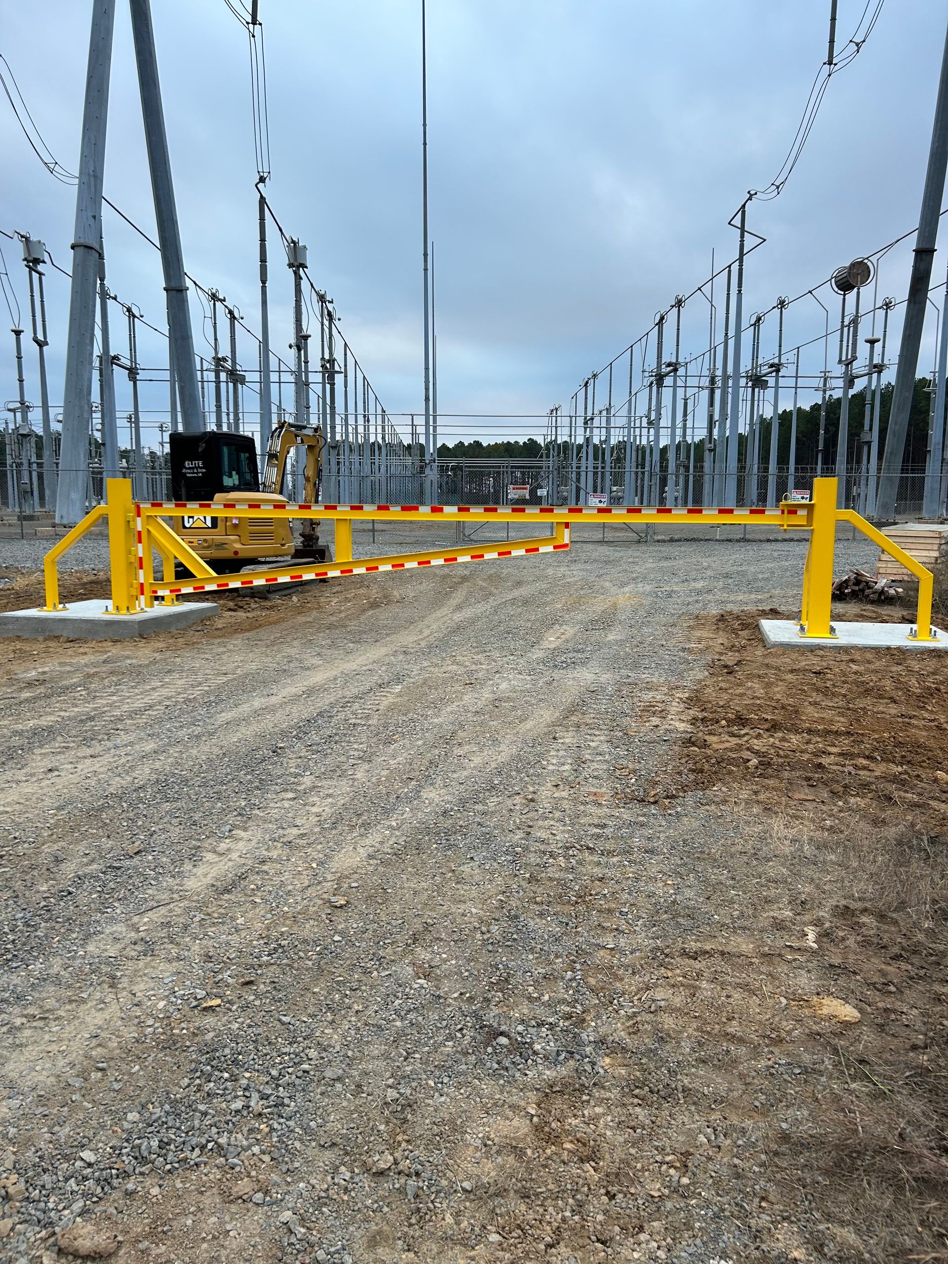 Yellow gate at an electrical substation. Excavator behind gate. Gray sky and gravel ground.