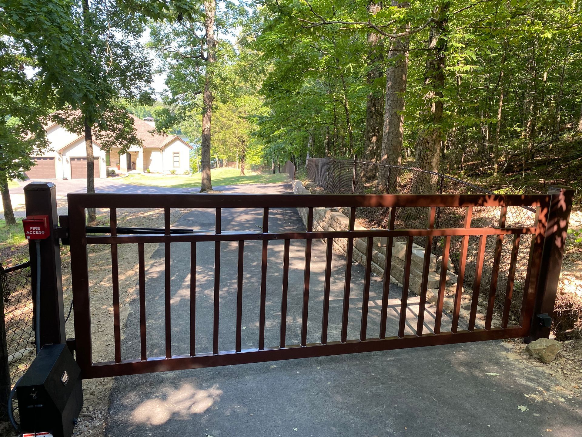 Brown metal gate at the entrance to a driveway. House visible in the background, surrounded by trees.