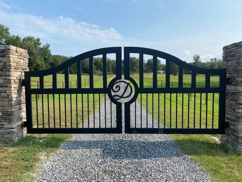 Black metal entrance gate with stone pillars, gravel driveway, and a grassy field in the background.