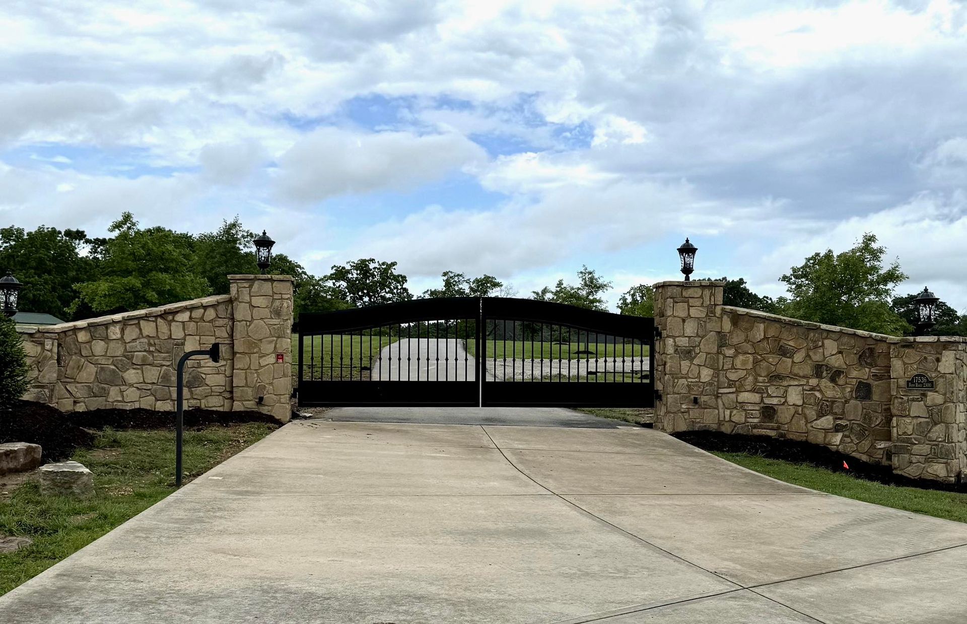 Stone pillars and black iron gate on a concrete driveway, under a cloudy sky.