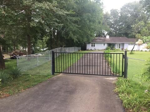 Black gate across a driveway leading to a white house, surrounded by trees and a chain-link fence.