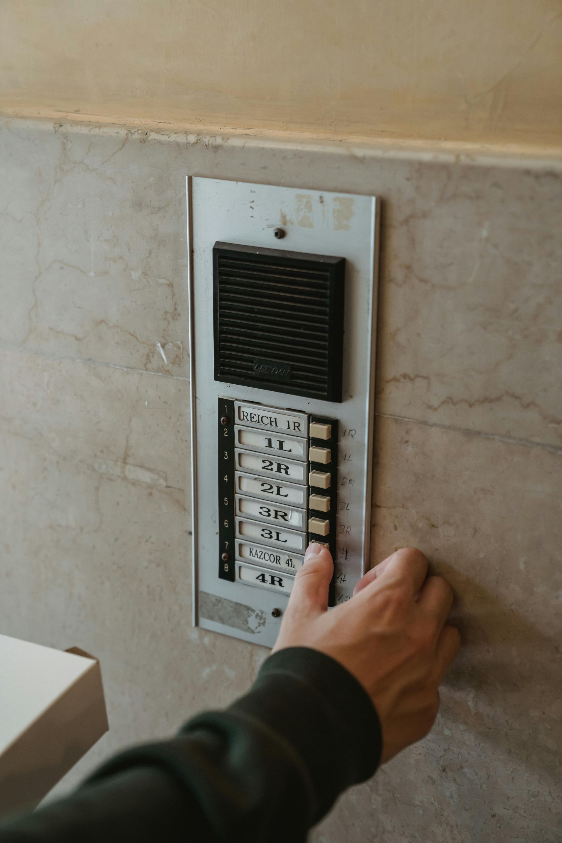 Hand pressing a button on a vertical intercom panel with black buttons, on a light-colored marbled wall.