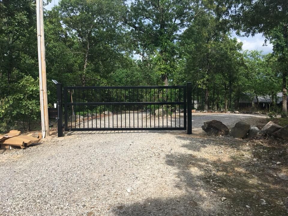 Black metal gate on a gravel driveway, leading to a wooded area.