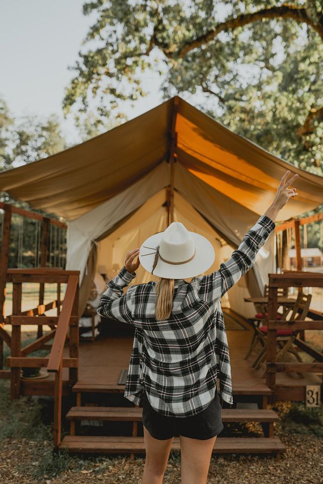 A woman in a hat is standing in front of a tent with her arms in the air.