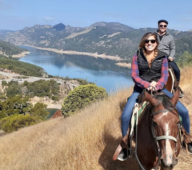 A man and a woman are riding horses on a hill overlooking a lake.