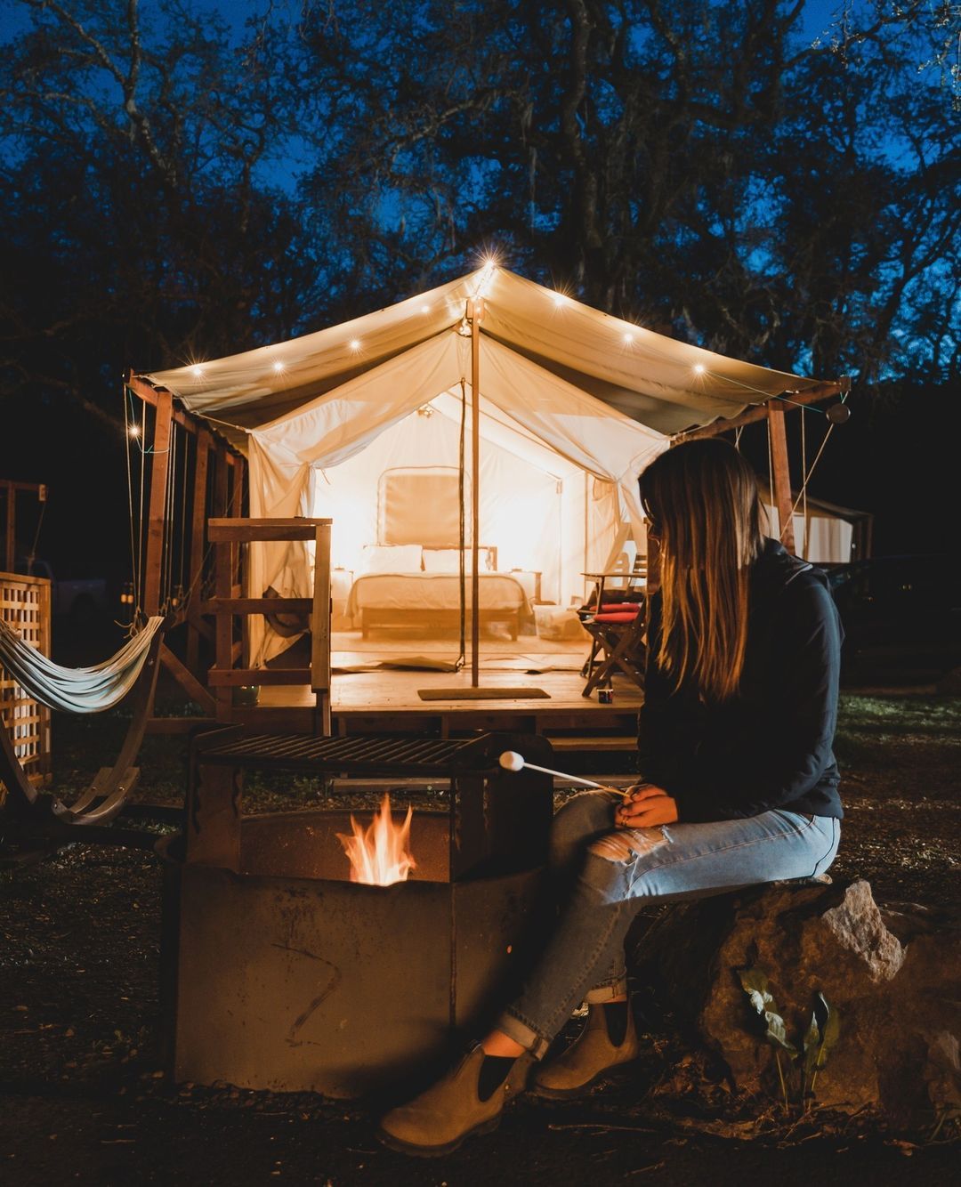 A woman is sitting next to a fire pit in front of a tent.
