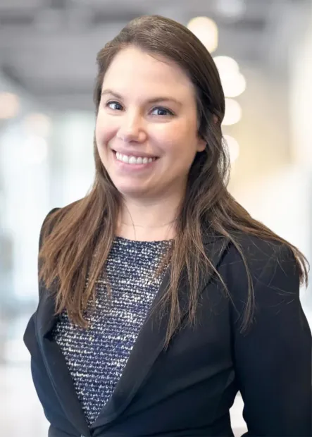 Woman smiling, wearing a black blazer and patterned top, in a bright office setting.