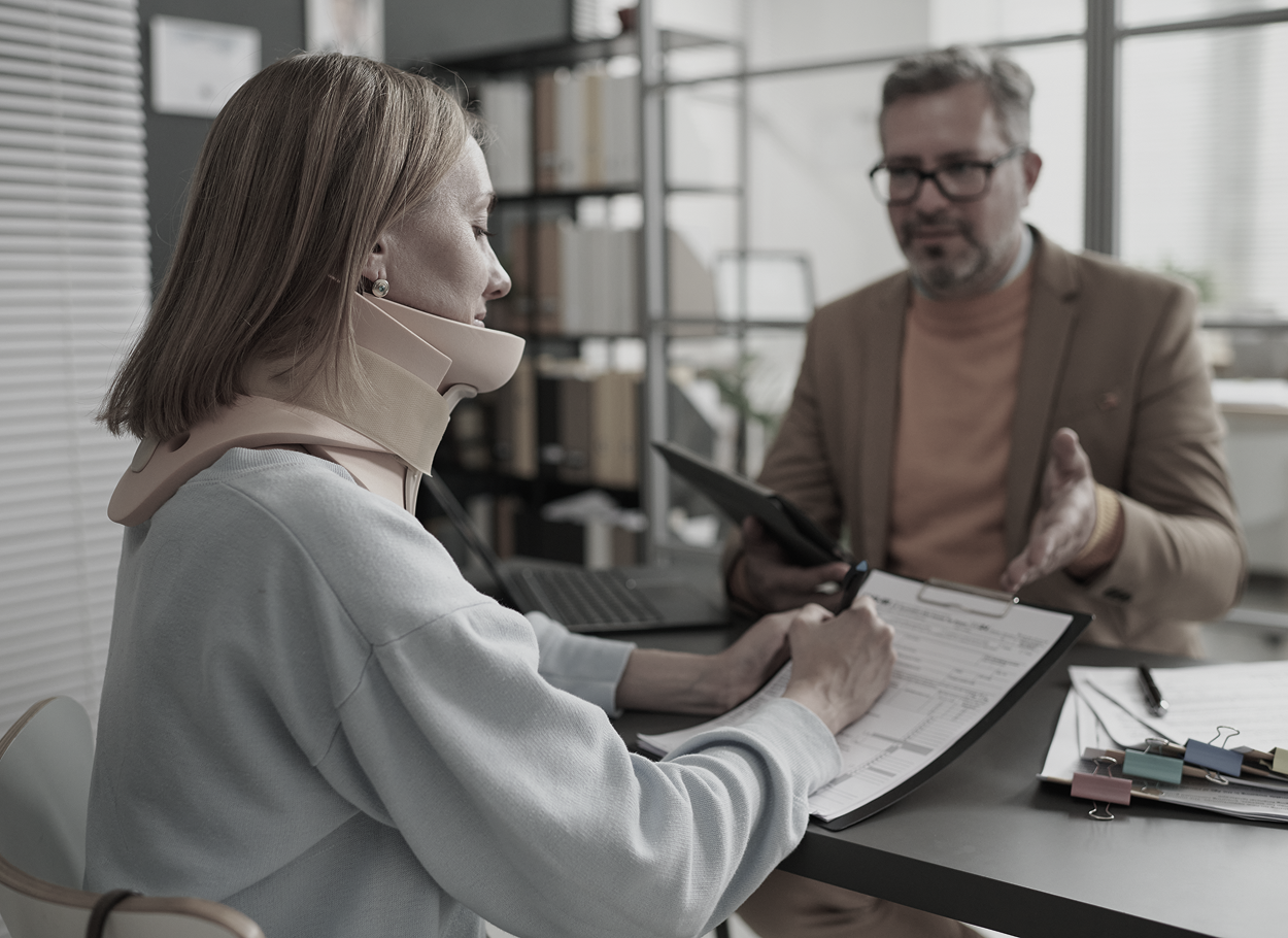Woman in neck brace signing documents at a desk while a man in glasses gestures, possibly in an office setting.