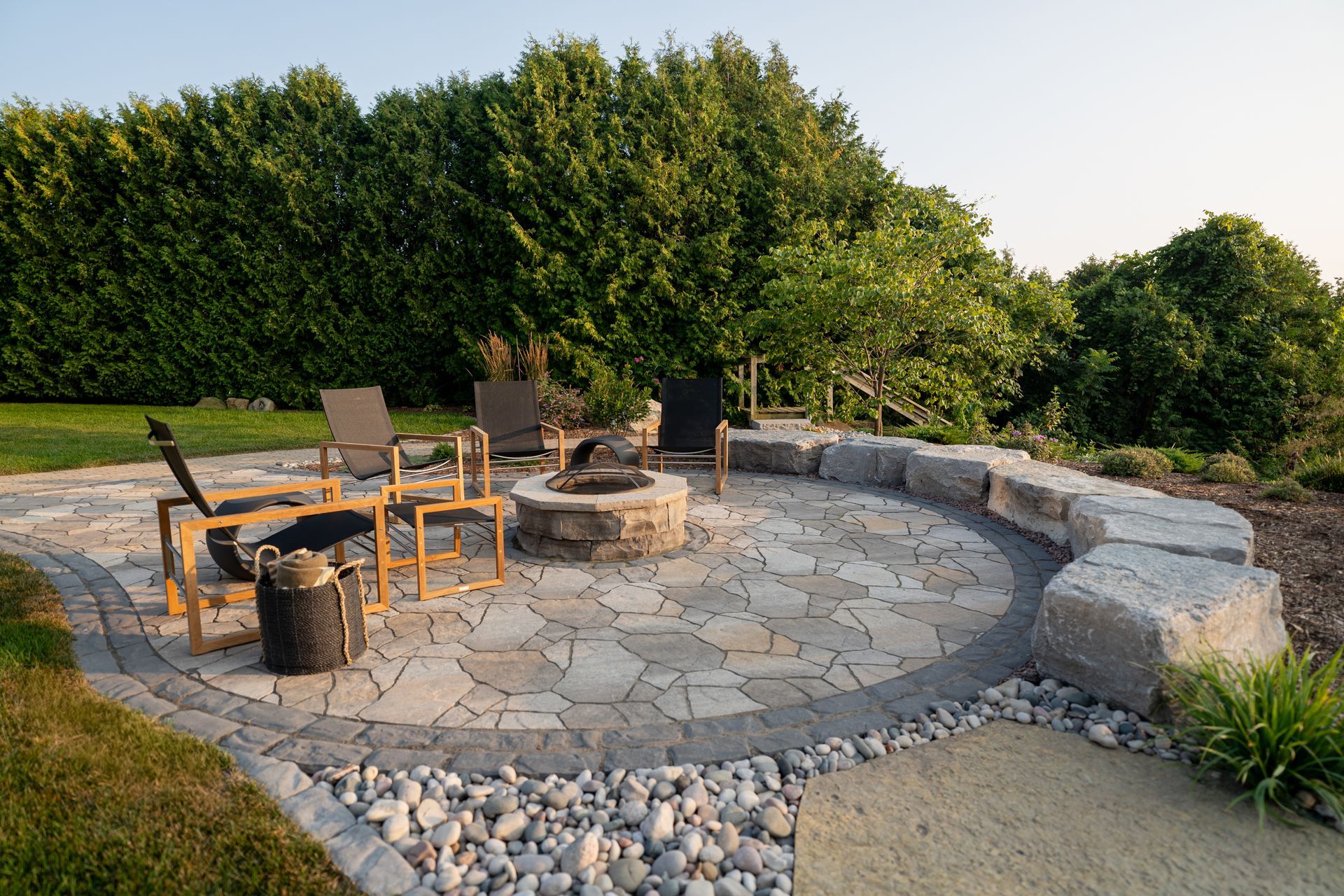 Stone patio with fire pit, seating, and landscaping; surrounded by greenery under sunlight.