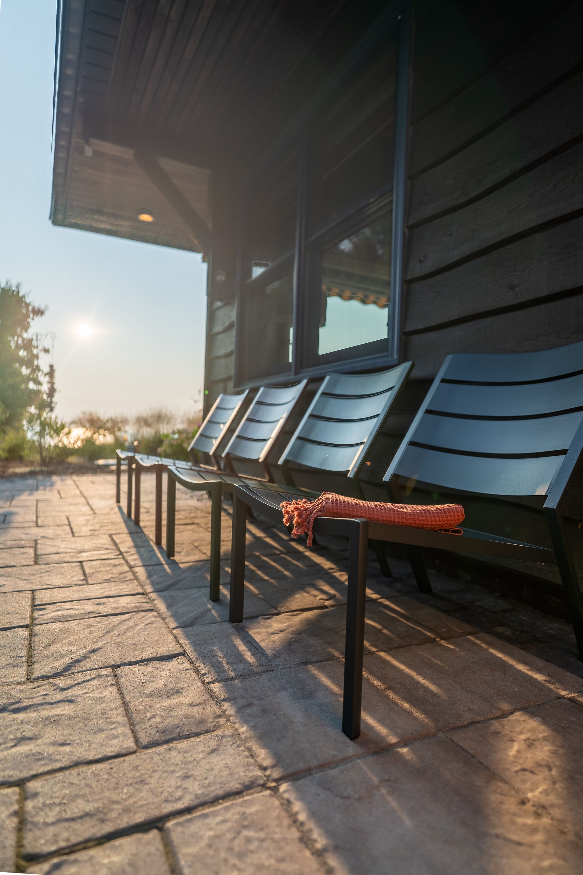 A row of black chairs on a brick patio with a wooden building in the background; sunlight.