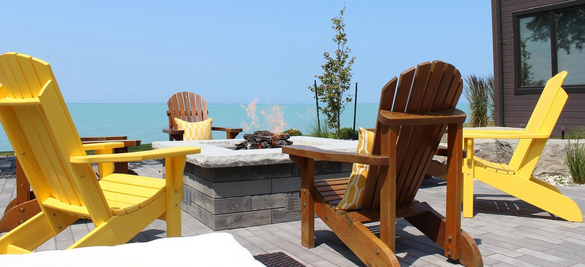 Adirondack chairs around a fire pit on a patio overlooking the water. Sunny day, yellow and brown chairs.