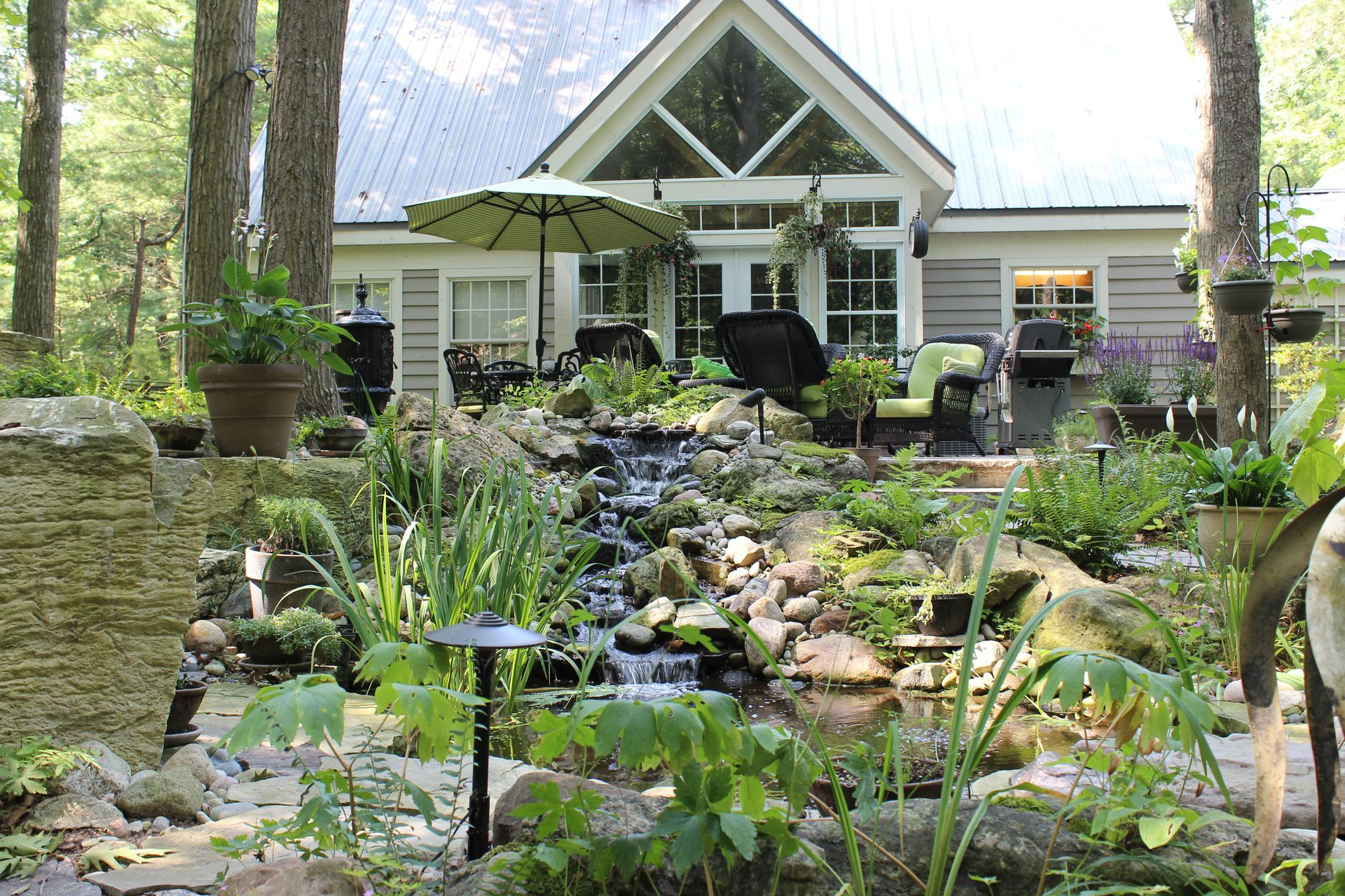 A house with a lush garden, waterfall, and pond. Green foliage surrounds the building with patio seating.