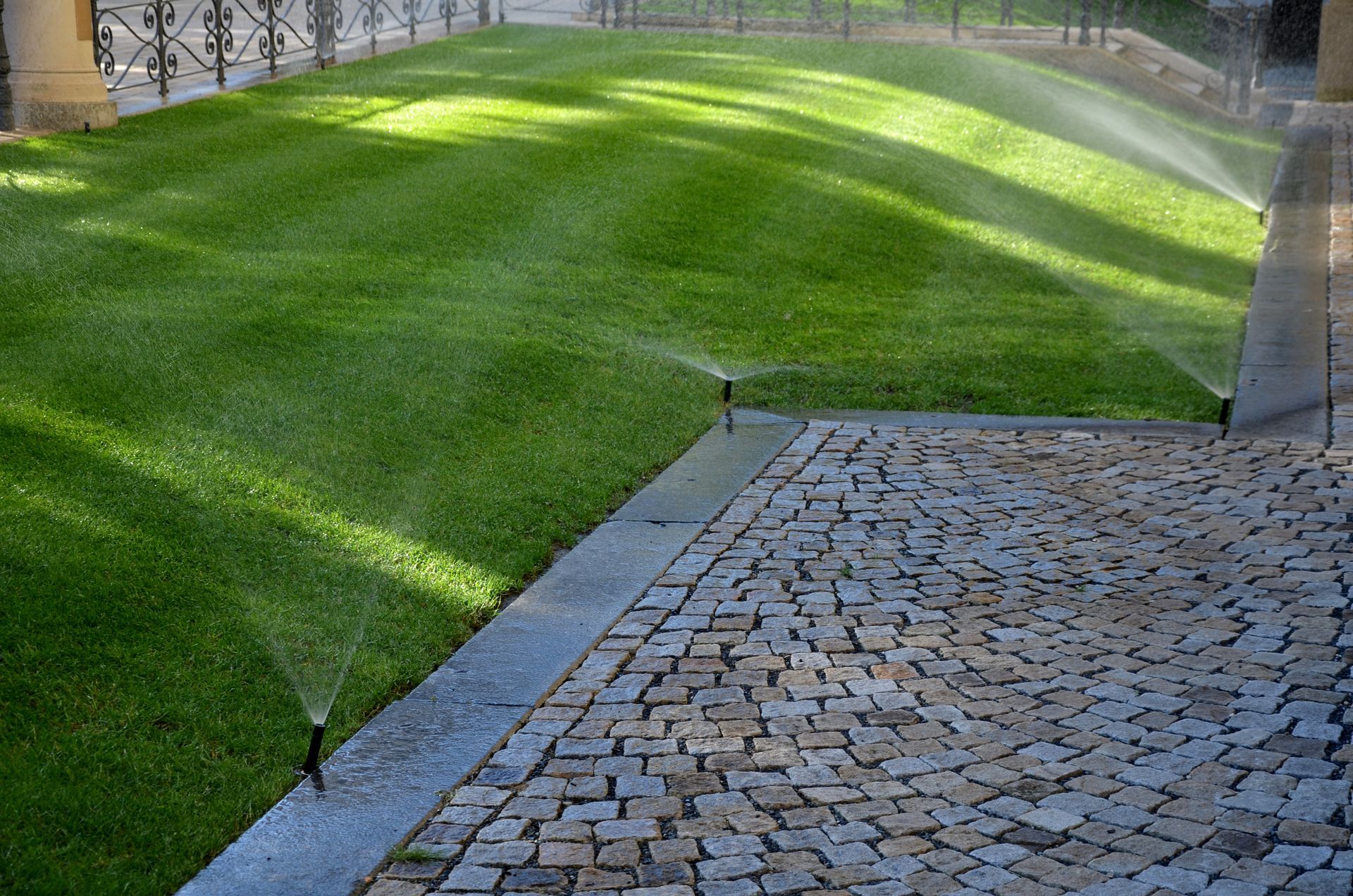 Sprinklers watering green lawn beside cobblestone walkway.