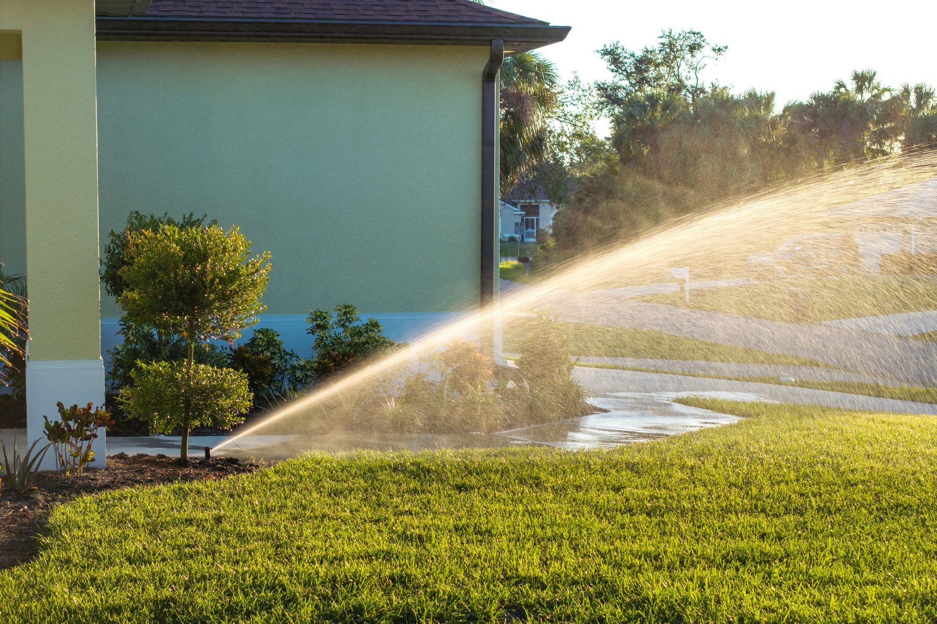 Lawn sprinkler spraying water over green grass in front of a light-green house.