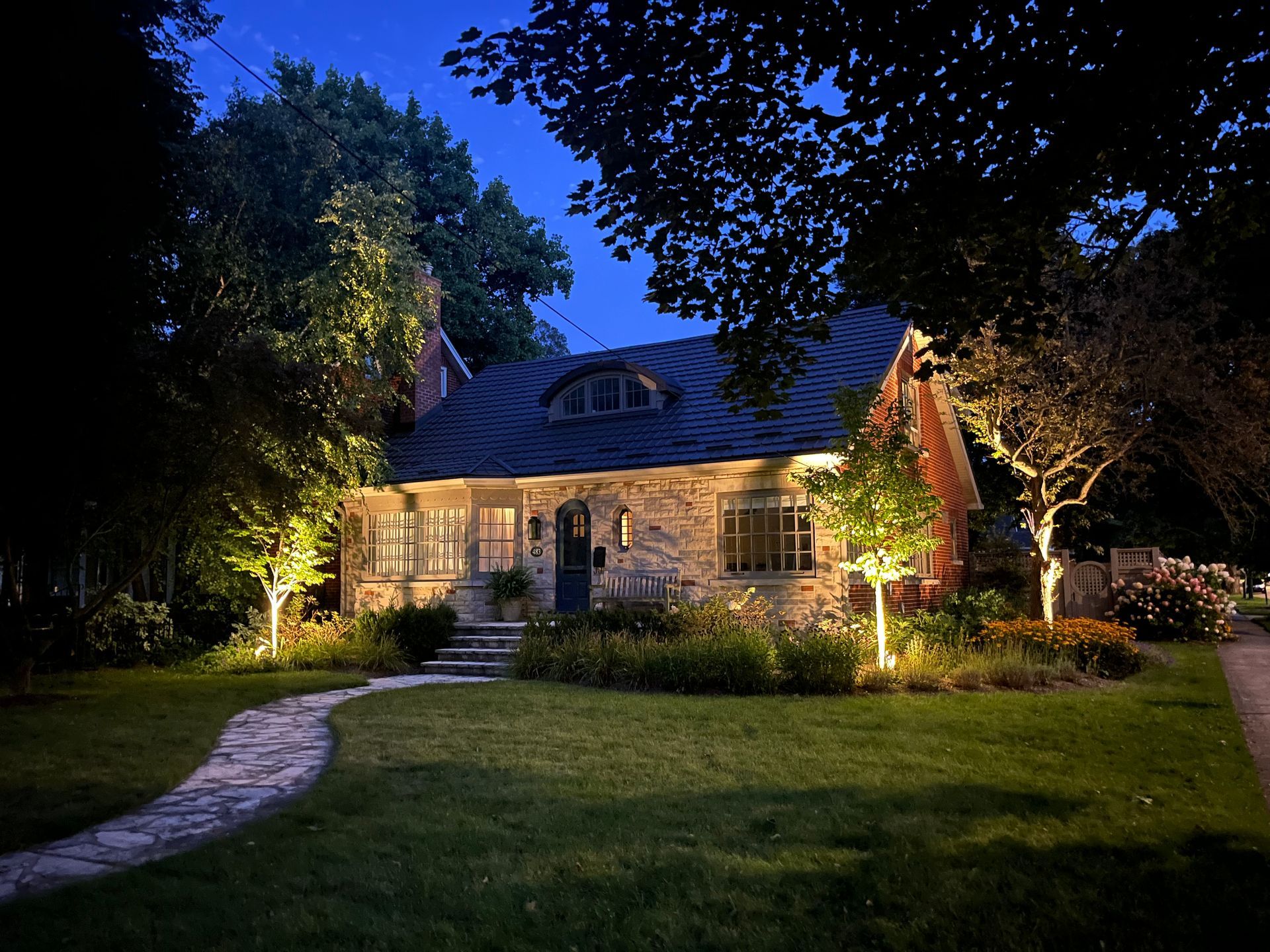 Stone cottage at night with illuminated trees and walkway. Dark blue sky.