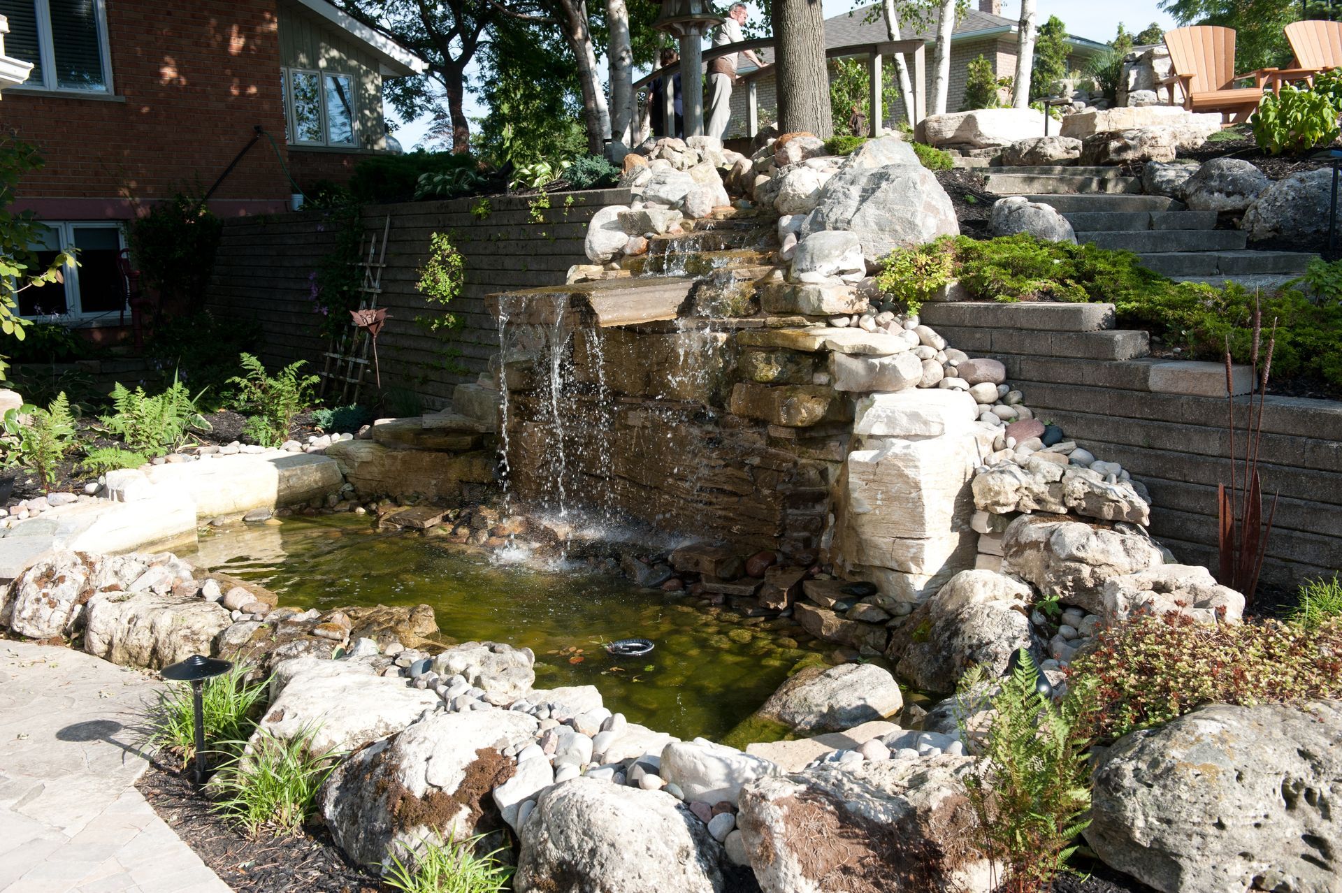 Water flows down a stone waterfall into a pond surrounded by rocks and greenery.