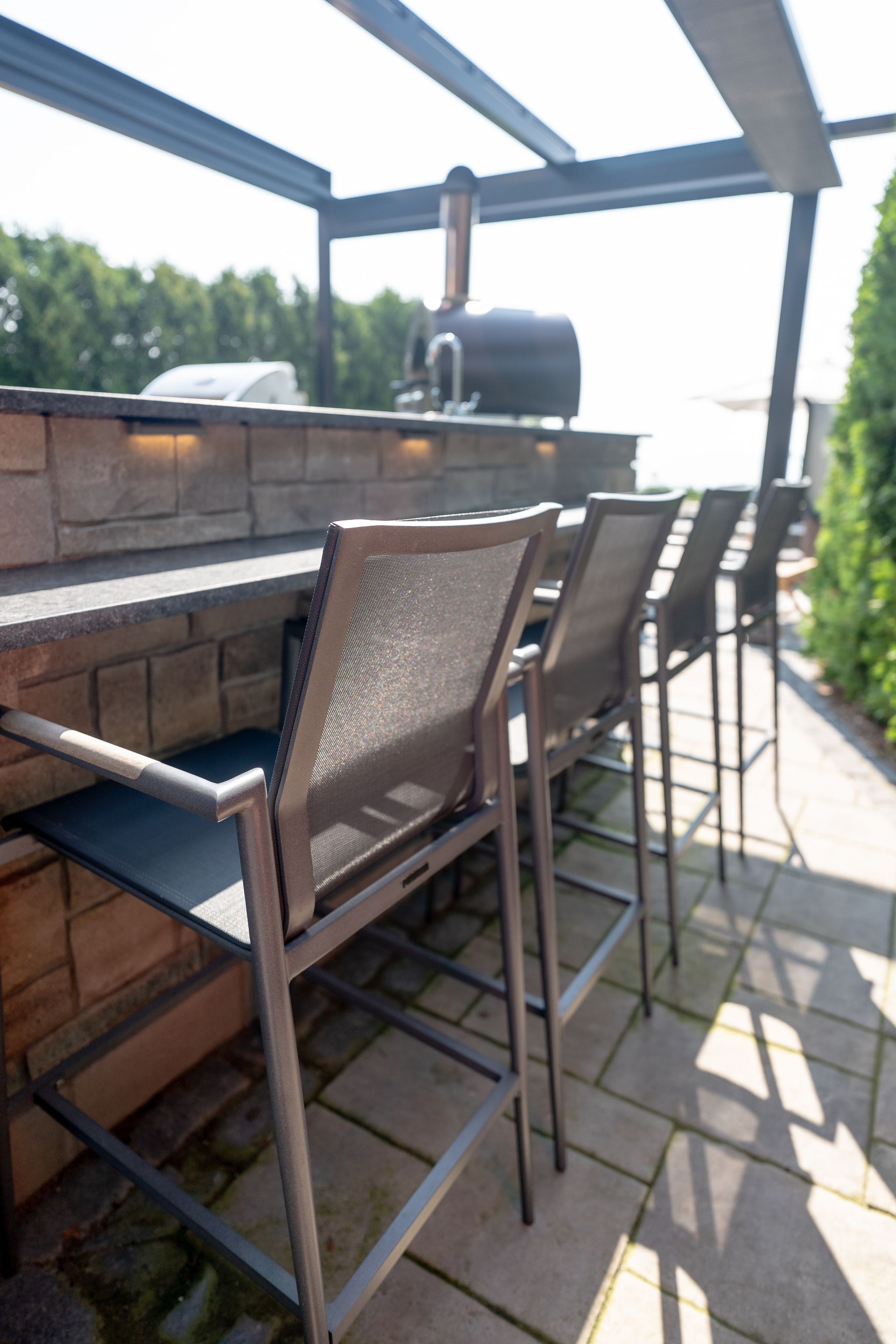 Outdoor bar with stools under a pergola. Brick-like counter, grill, and pizza oven.
