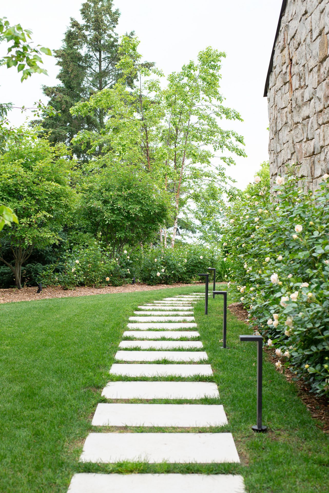 Stone path through green grass, lined with lights and flowering bushes, next to a stone building.