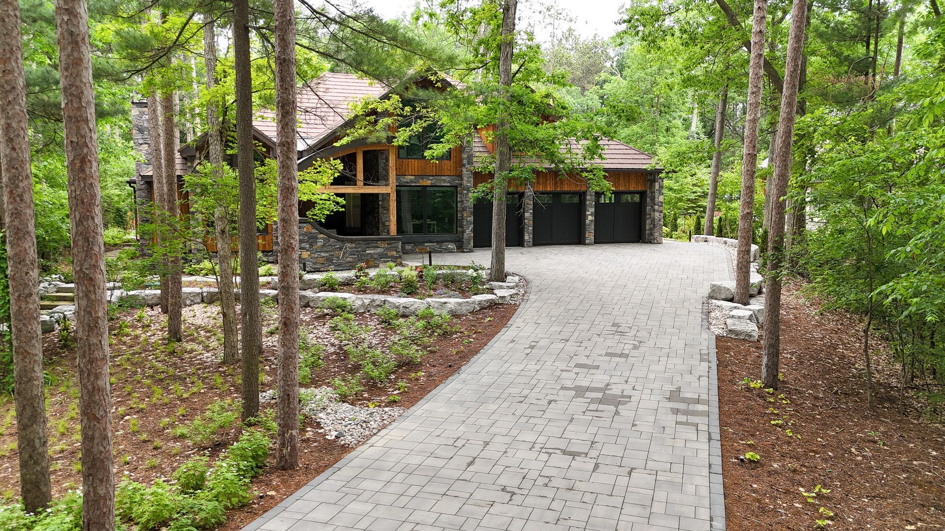 Stone and wood home with a brick driveway surrounded by trees.