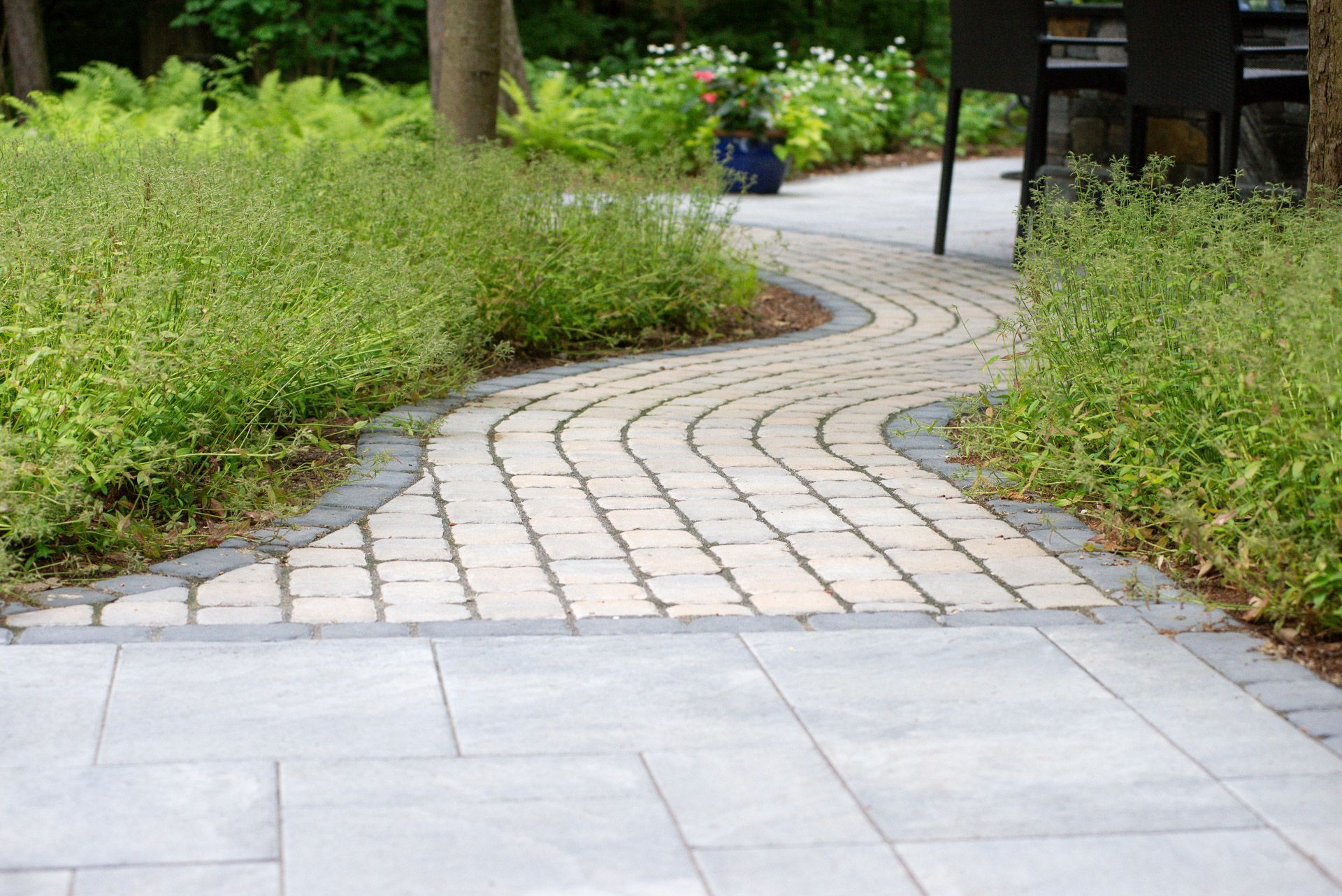 Winding brick pathway through a garden, bordered by green bushes. Blue-gray stone pathway leads to it.