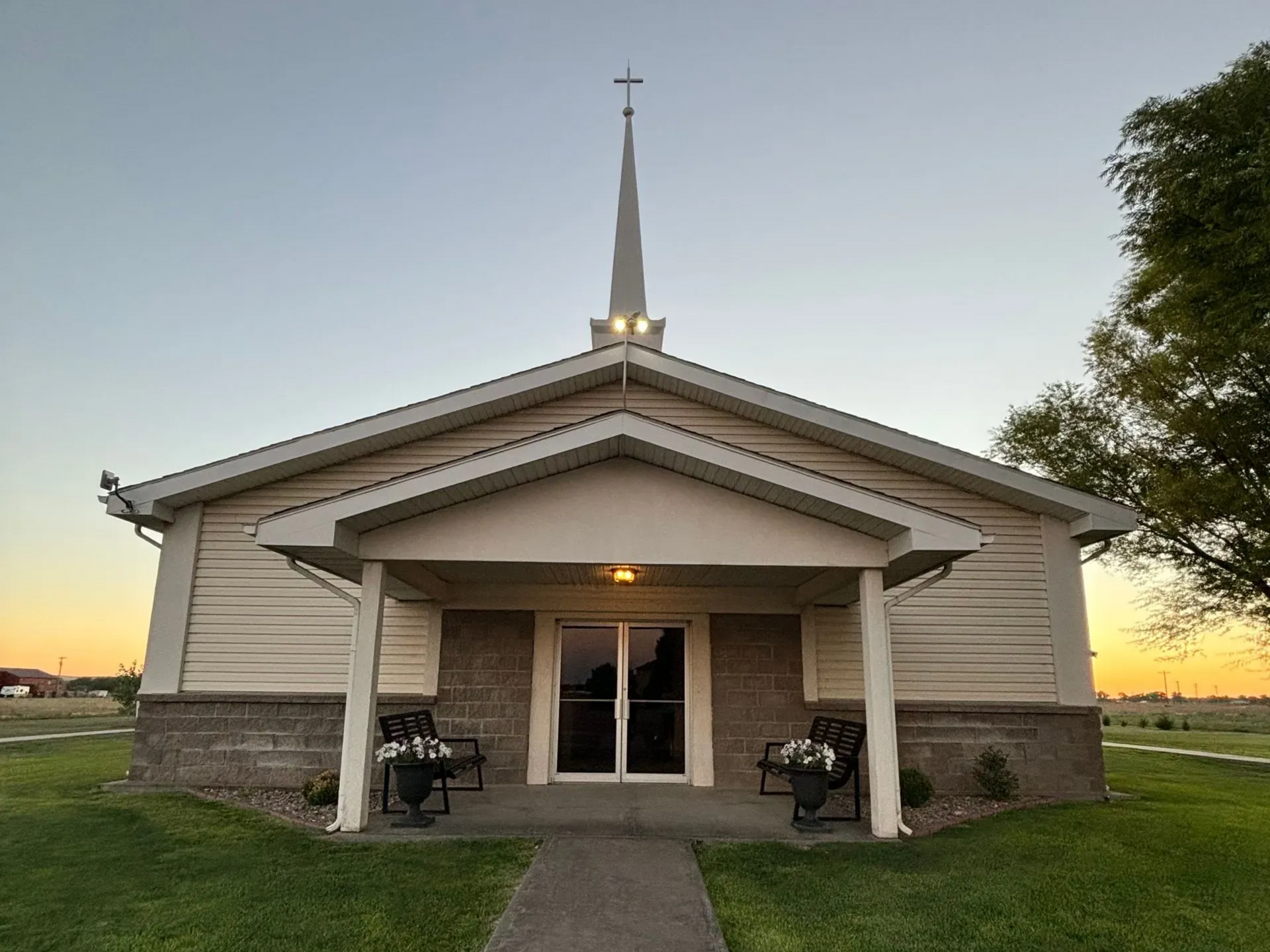 Small white church with a steeple at sunset, framed by trees and lawn.
