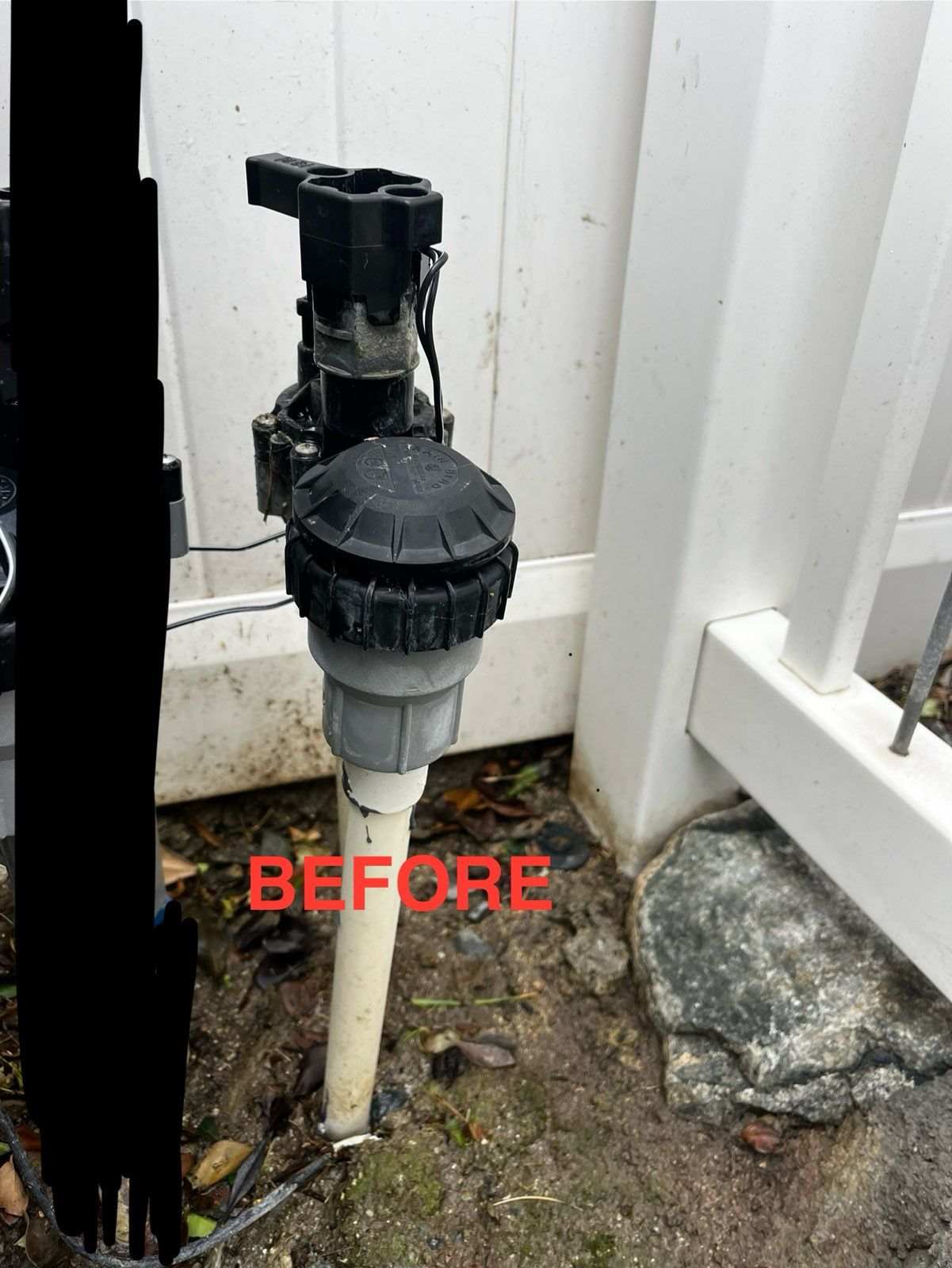 A sprinkler valve is sitting on the ground next to a white fence.