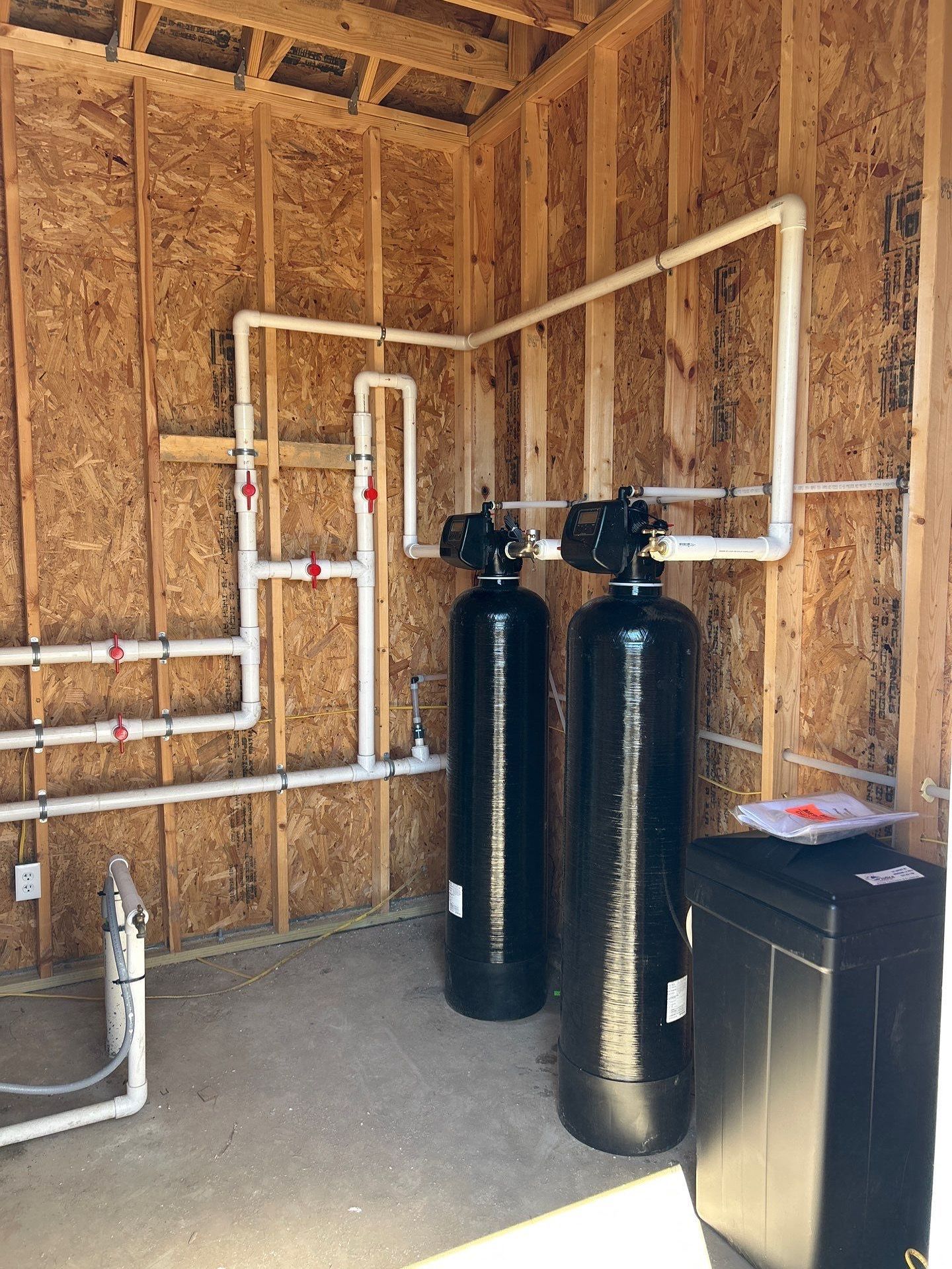 Water filtration system installed against a concrete wall, with stainless steel tanks and blue filters, copper pipes.