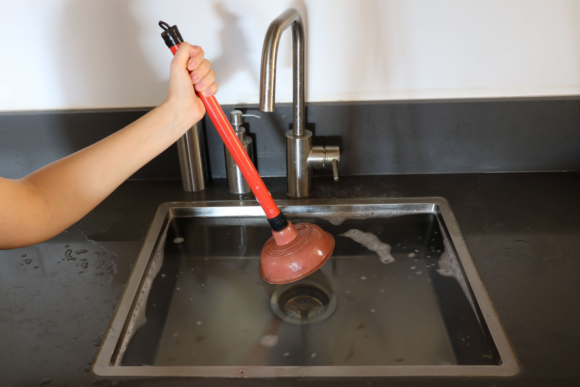 A person using a red plunger in a stainless steel kitchen sink to unclog a drain.