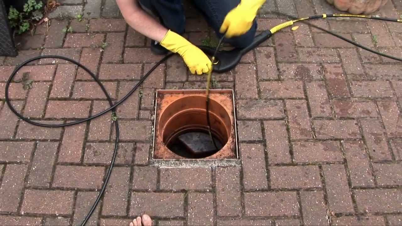 Person wearing yellow gloves, cleaning a drain, on a brick patio.