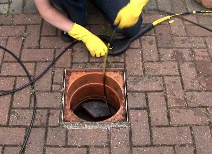 Workers inspecting a sewer line, using tools near an open manhole on a paved road.