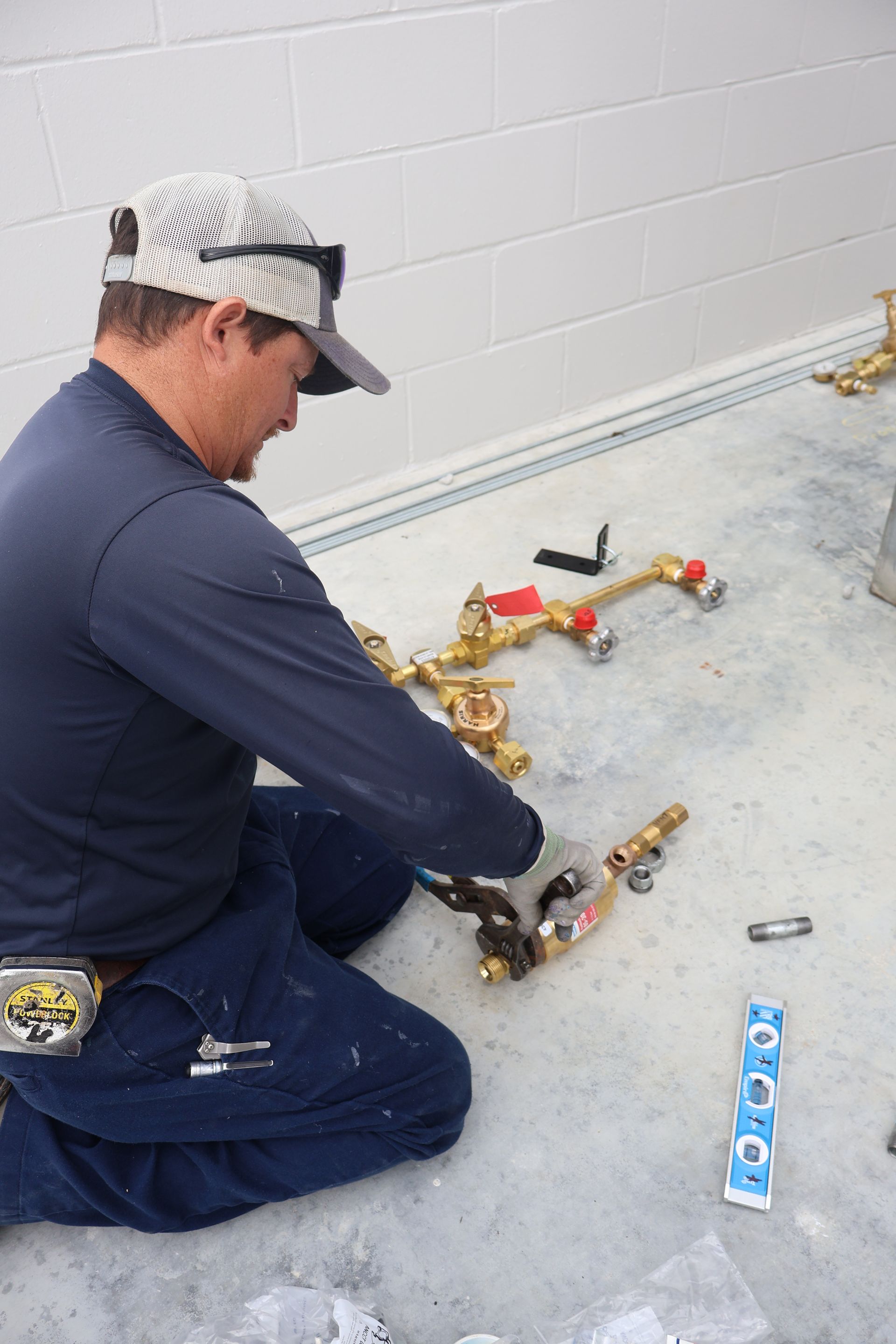 Plumber sits on floor, assembling brass plumbing fixtures. Gray concrete floor, white wall, blue shirt.