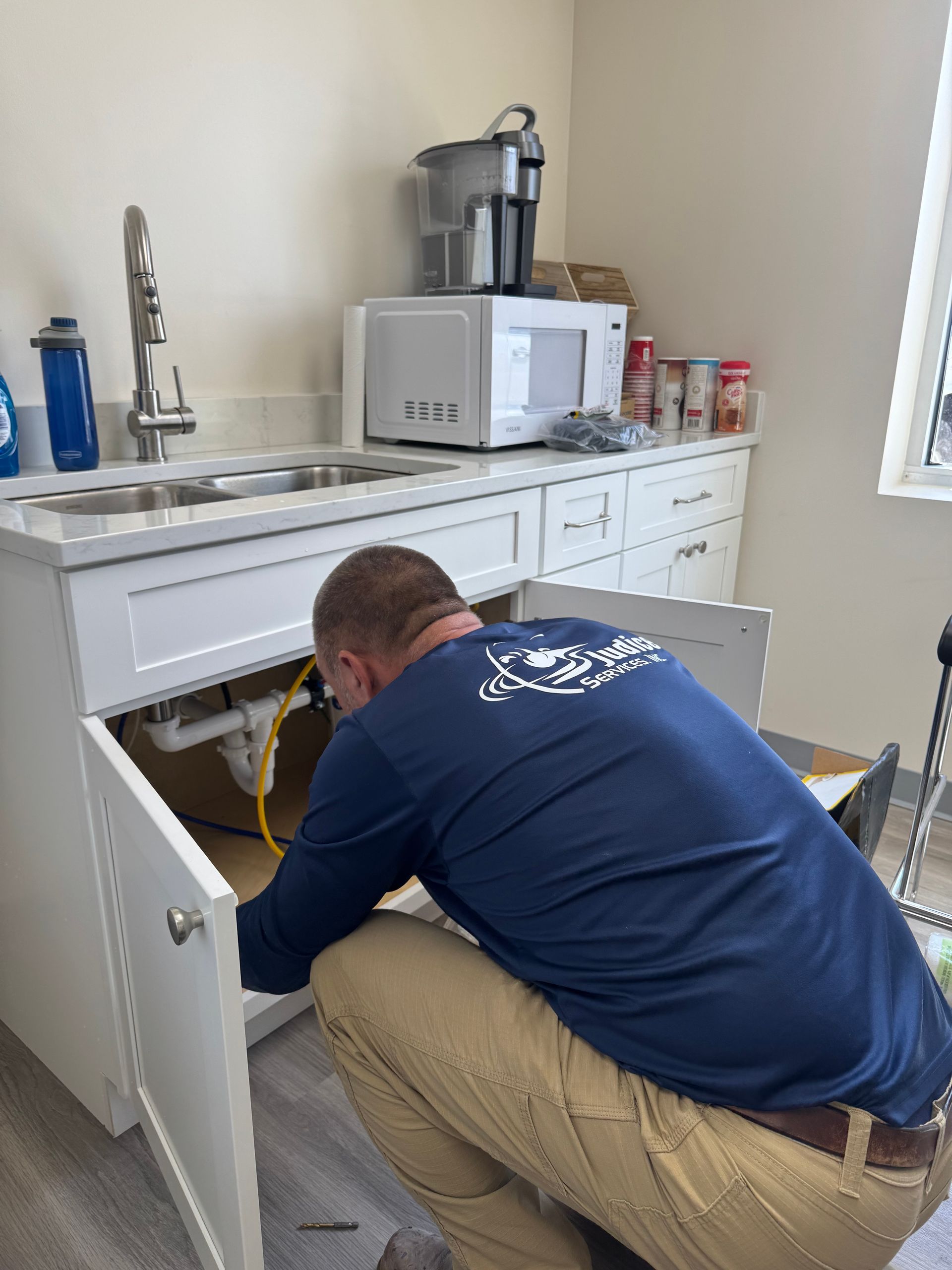 Man in blue shirt working under a white sink in a kitchen.