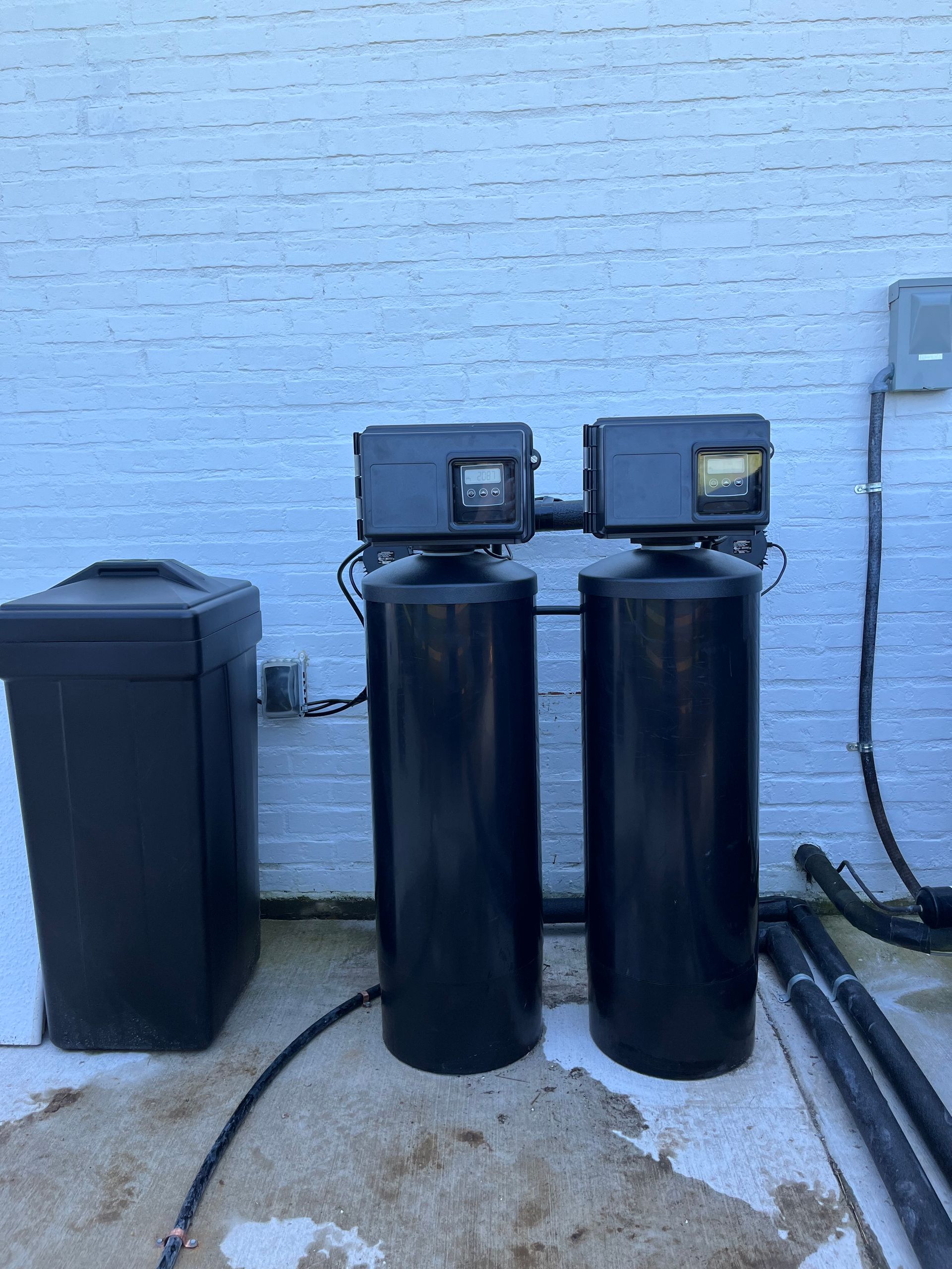 Two black water filtration tanks with control panels next to a salt storage bin, outside against a white brick wall.