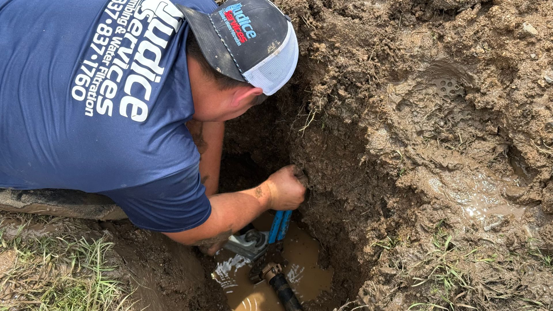 A plumber in a blue shirt repairs a pipe in a muddy hole.