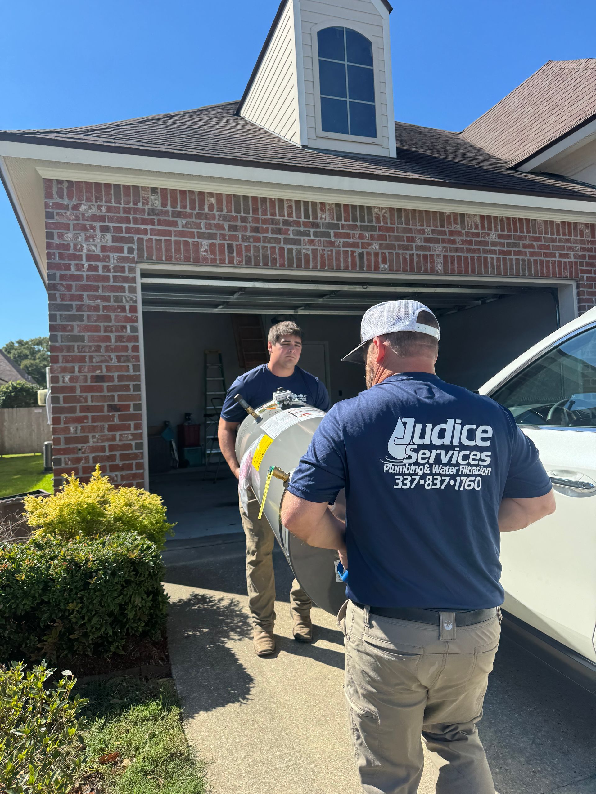 Two men in blue shirts carrying a cylindrical object into a garage next to a white vehicle.