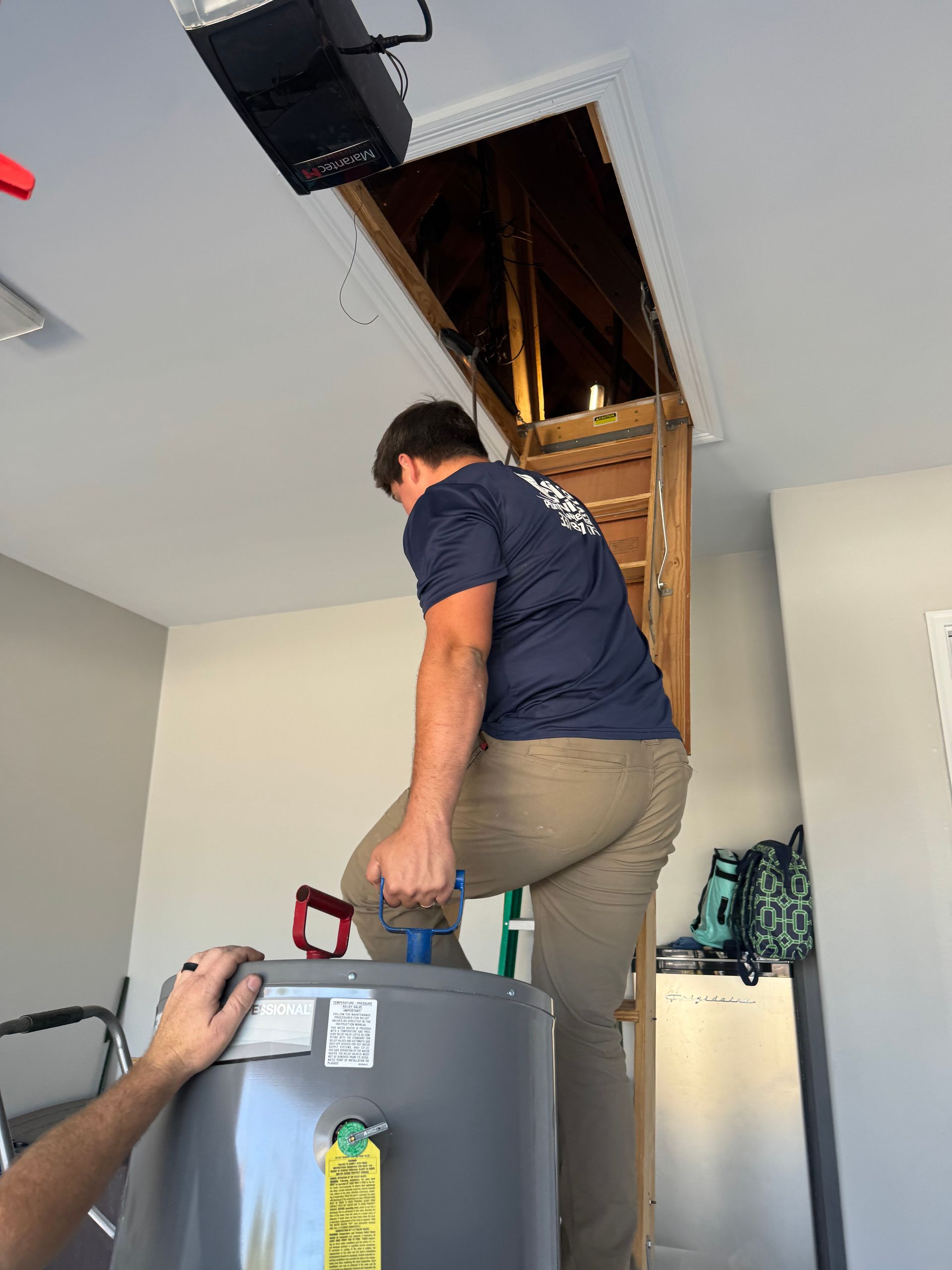 Man climbing a pull-down attic ladder carrying a water heater, person below helping in a garage.