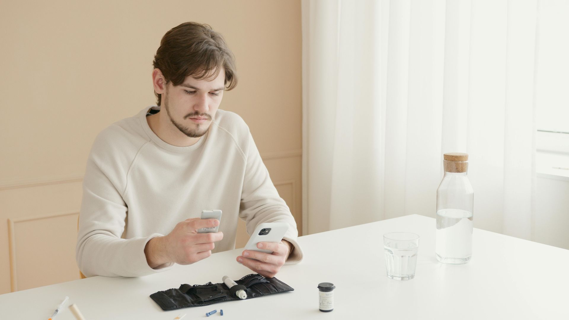 Man sitting at a white table, looking at phone, holding glass, and insulin testing kit in front of him.