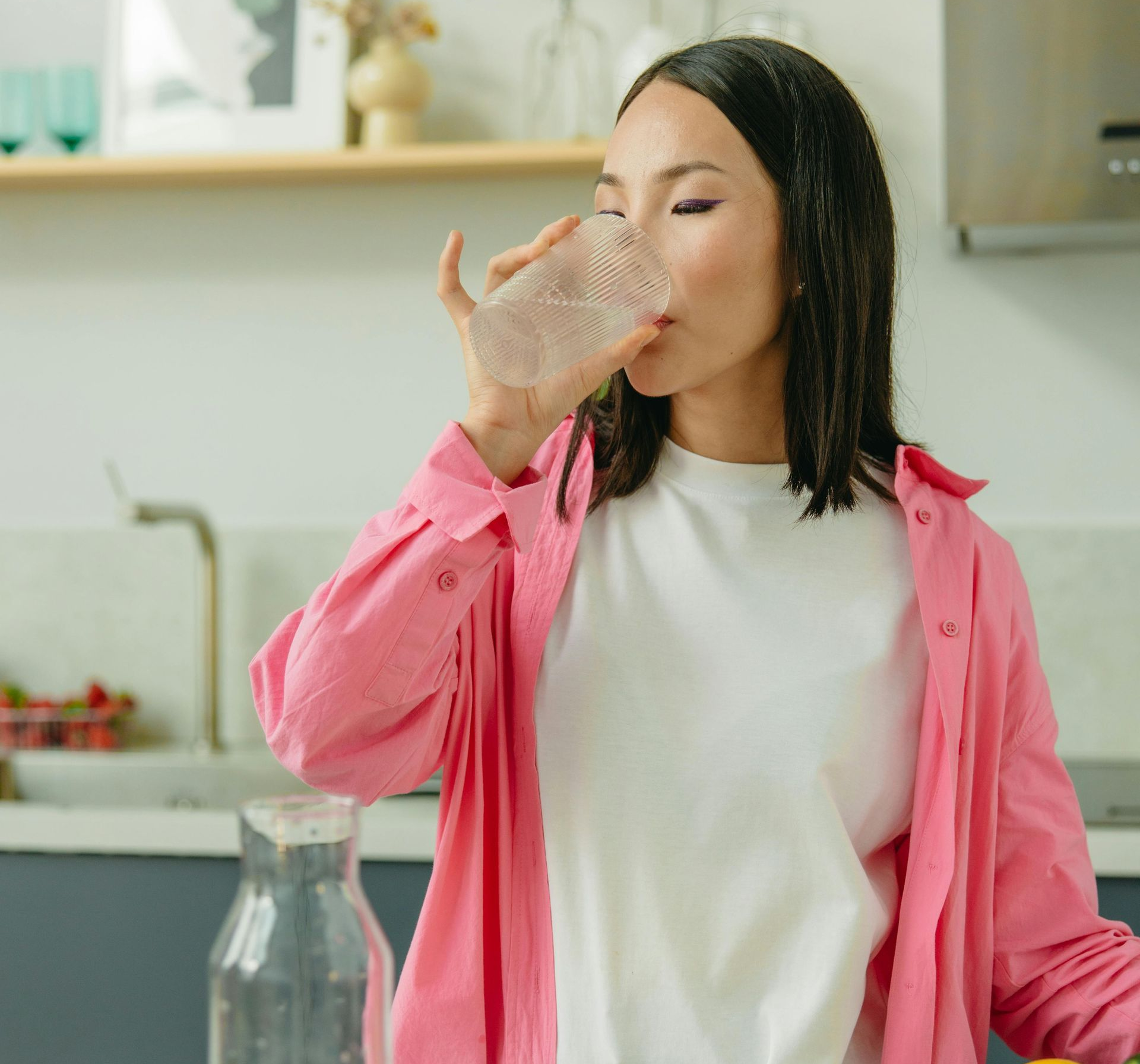 Woman drinking from a glass in a kitchen, wearing a pink shirt over a white tee.