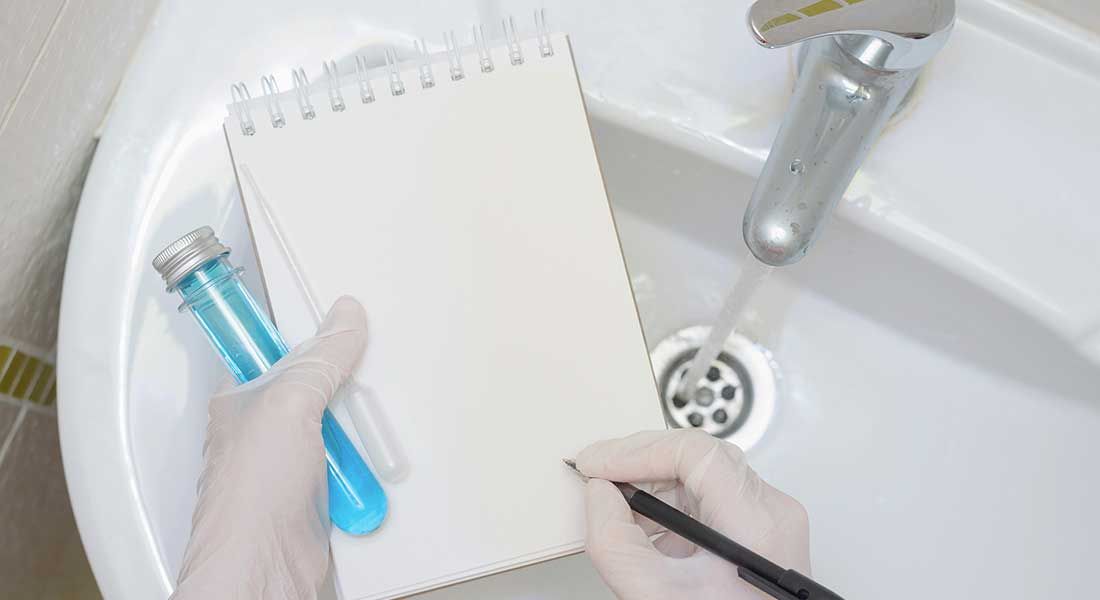 Person with gloves writing in a notebook near a running faucet and blue test tube in a sink.