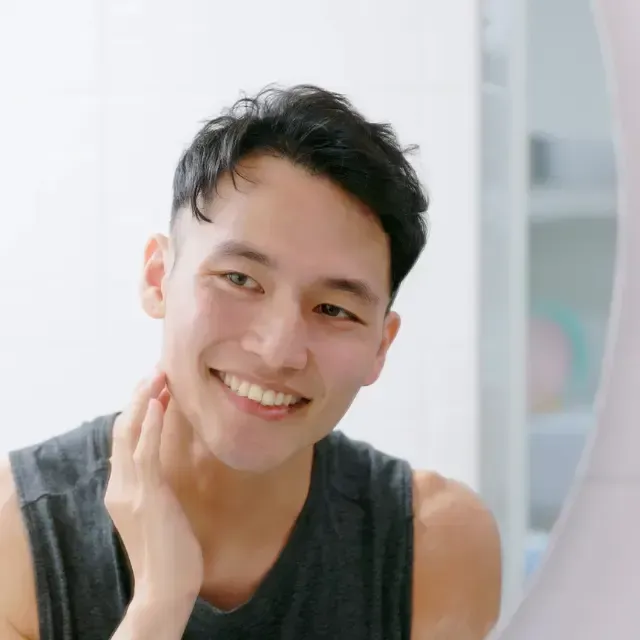 A man is smiling and touching his face in front of a mirror.
