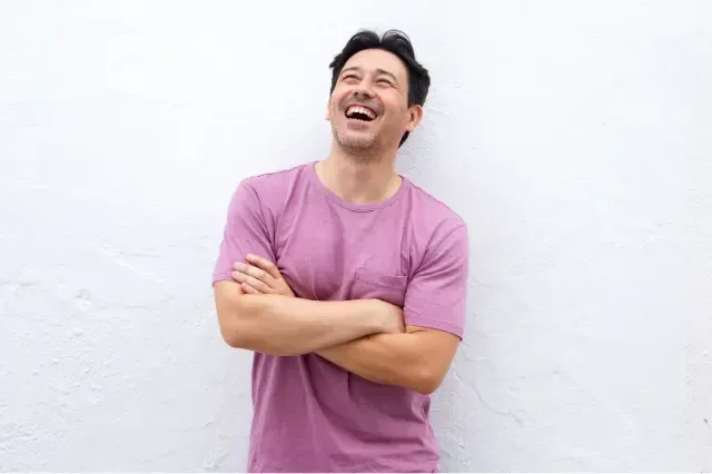 A man in a purple shirt is standing in front of a white wall with his arms crossed and laughing.