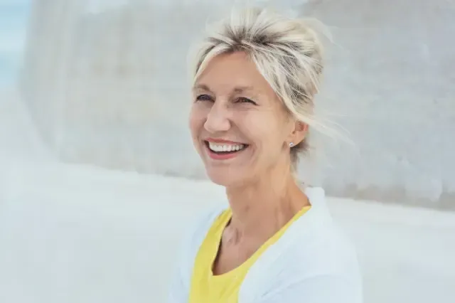 An older woman is smiling while sitting on a beach.