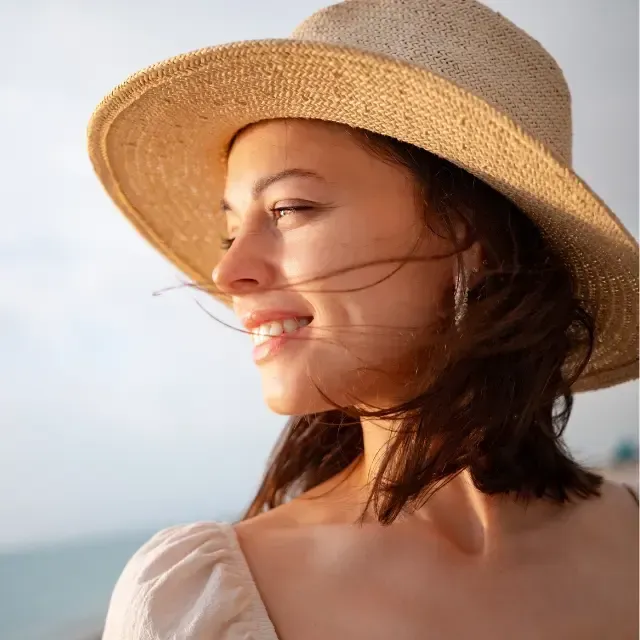 A woman wearing a straw hat is smiling with her hair blowing in the wind