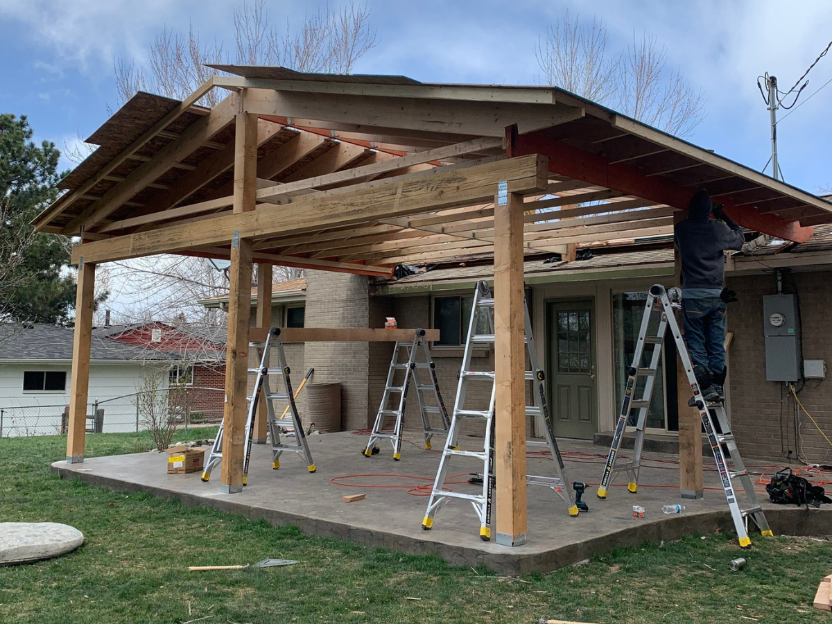 A person stands on a ladder working on the wooden frame of a patio cover attached to the back of a brick house.