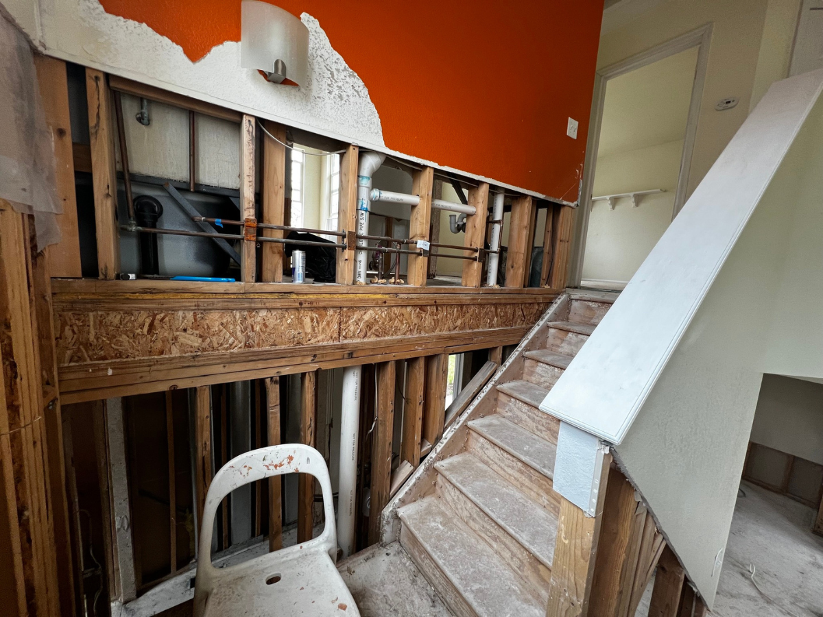A home renovation in progress with exposed wooden wall studs, plumbing pipes, orange paint, and a wooden staircase.