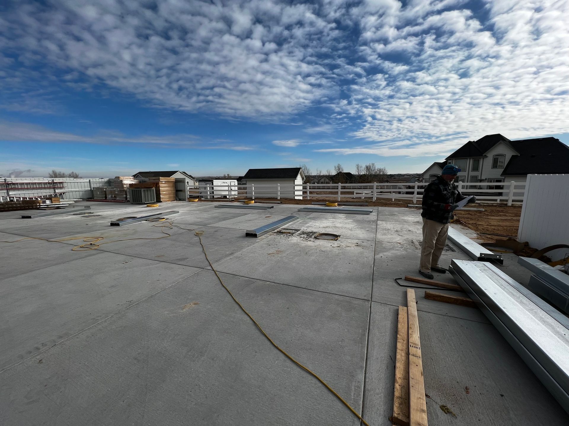A person in a hard hat inspects a concrete building foundation at a construction site under a partly cloudy blue sky.